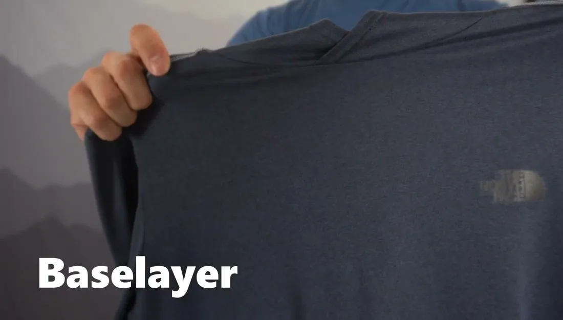 A guide's hands holding up a long-sleeve dark blue-gray base layer shirt in front of a mountain-print backdrop, with the word Baselayer on screen.