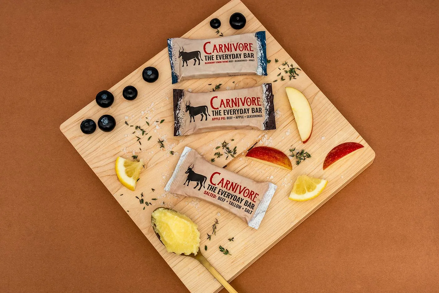 Three Carnivore Bar Everyday Bars in Blueberry Lemon Thyme, Apple Pie and Salted on a wooden board with a spoon of tallow, apple slices, lemon and blueberries