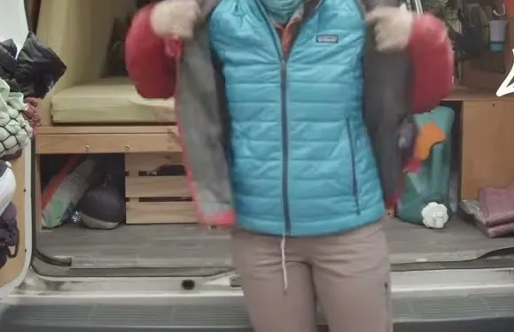A hiker standing behind an open van, pulling a red hard shell jacket on over a blue quilted puffy vest.