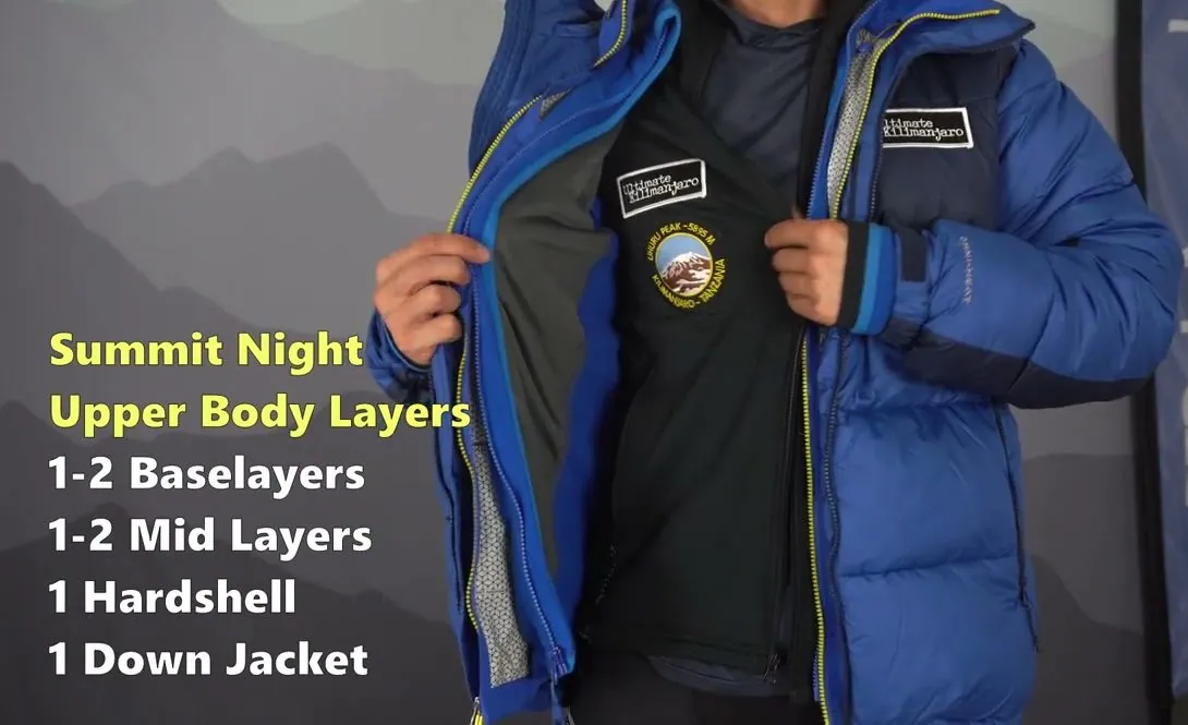 A guide opening a blue down jacket to show the dark mid layer and base layer underneath, with an on-screen list: Summit Night Upper Body Layers, 1 to 2 baselayers, 1 to 2 mid layers, 1 hardshell, 1 down jacket.