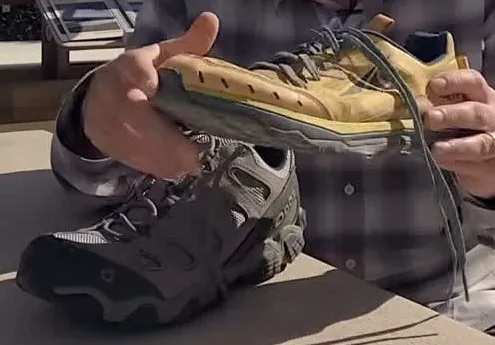 A hand holding a yellow trail runner shoe above a gray hiking shoe with a thick tread, both on a table outdoors.