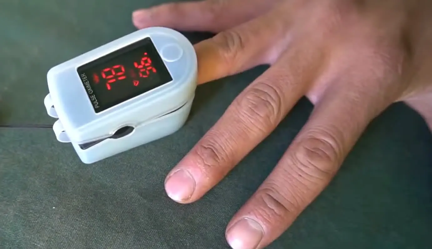 A white fingertip pulse oximeter with a red digital display clipped on a finger of a hand resting on a green canvas surface.