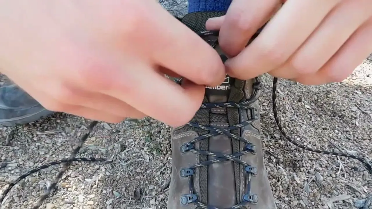 Two hands pinching the crossed laces at the top eyelets of a brown hiking boot laced up to the ankle