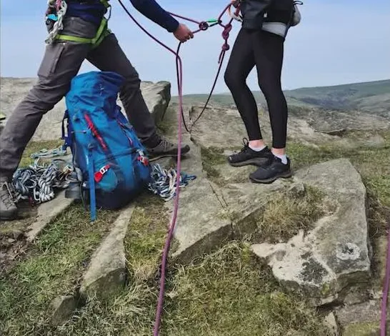 Two people on a rocky hilltop working with a purple rope. A blue backpack and coiled climbing gear sit on the rock between them, and one person wears a harness.