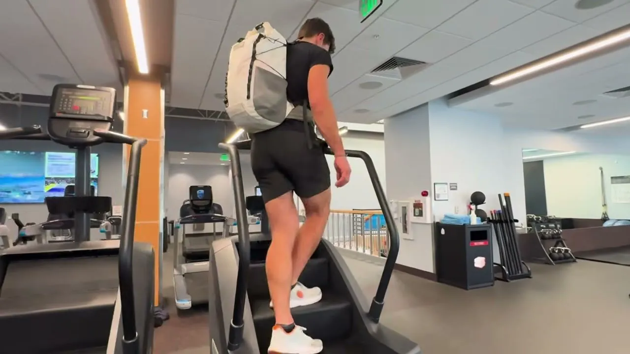 A person seen from behind climbing a stair machine in a gym while wearing a white backpack.