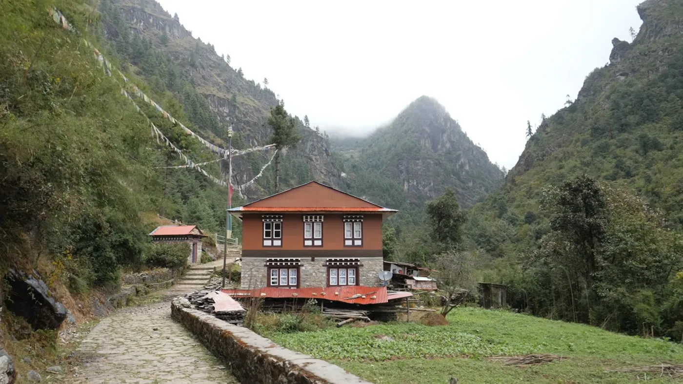 A two-story guesthouse with a red roof and small windows beside a stone path in a green, misty valley, with prayer flags strung across the trail.