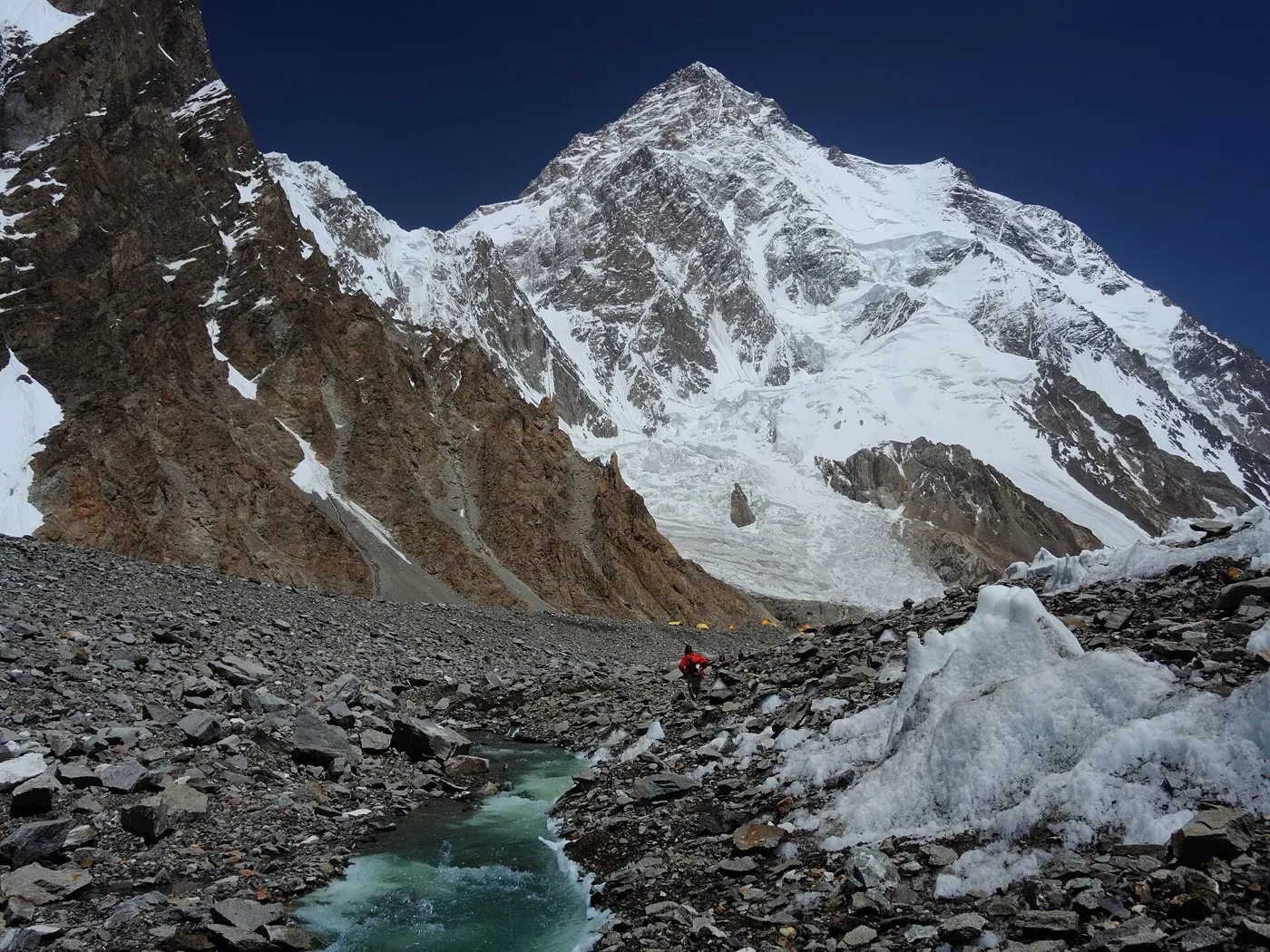 A rubble-covered glacier with a green meltwater stream and a small hiker in red, base camp tents tiny at the foot of K2's south face
