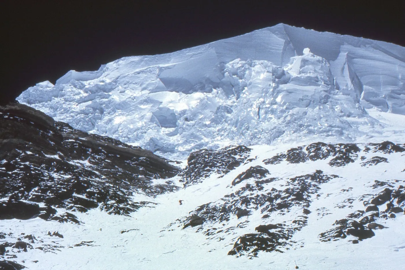 A huge broken ice cliff on K2 above a narrow snow gully between dark rock bands, with one tiny climber in the gully