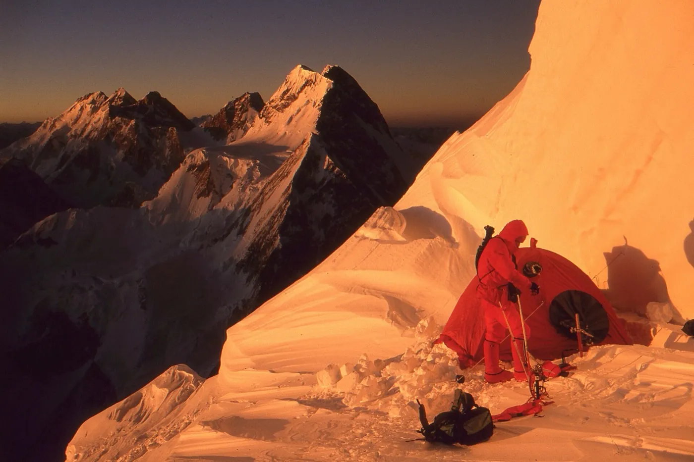 A climber in a red down suit beside a red tent dug into a steep snow slope at sunrise, with orange-lit peaks behind