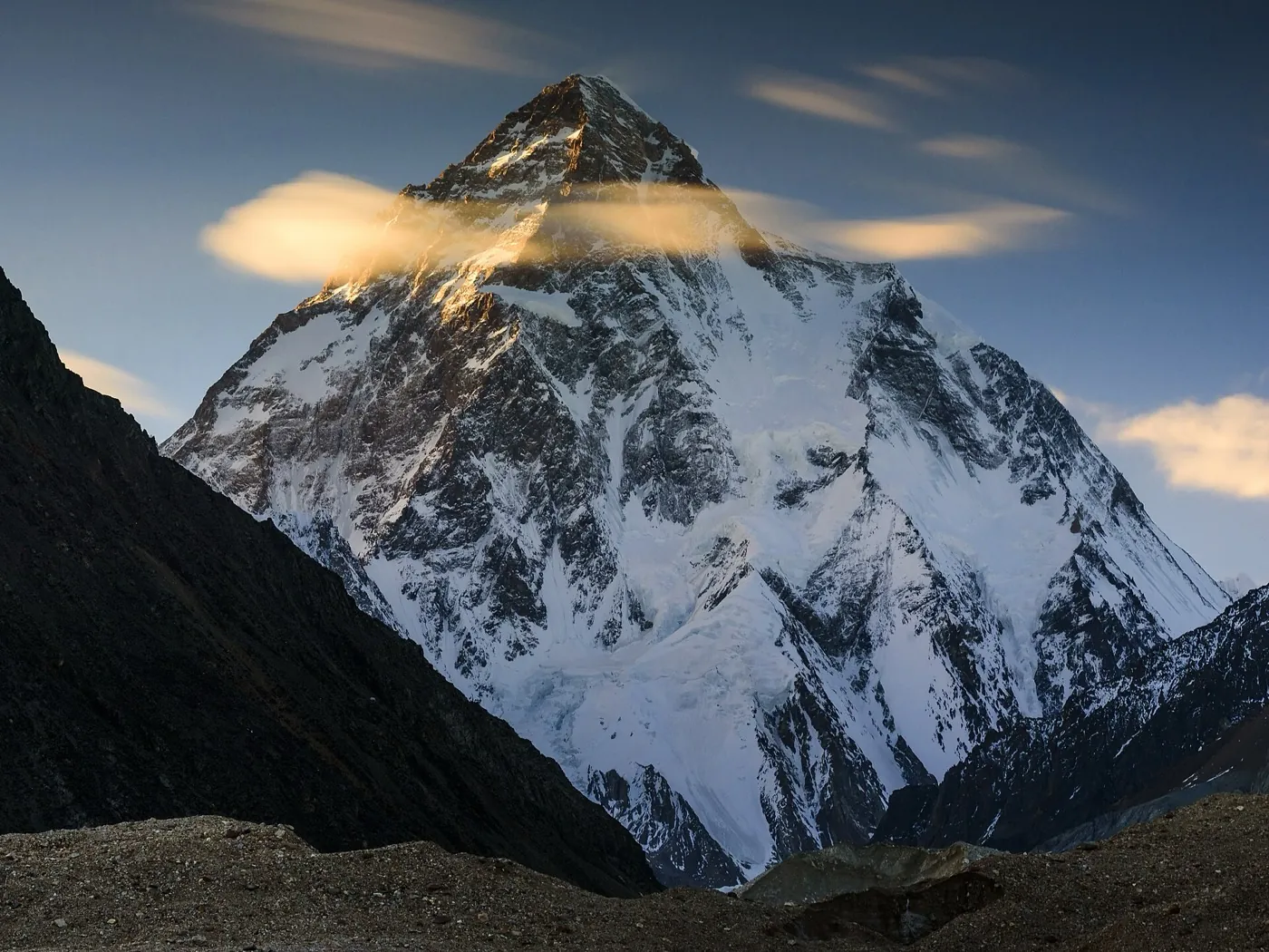 K2's south side at dusk, a steep snow and rock pyramid with its summit lit and small clouds around it, dark moraine in front