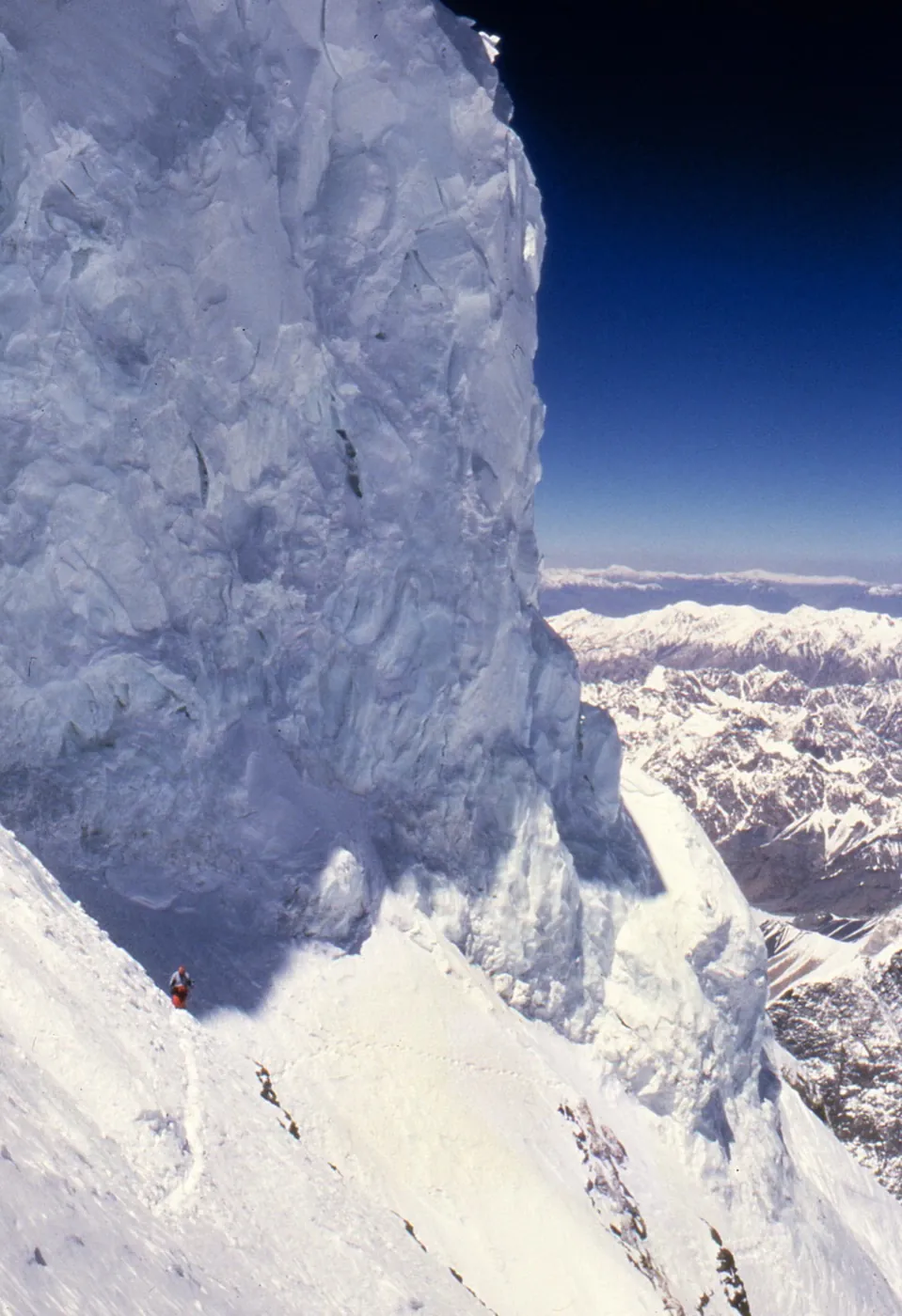 A climber in a red suit on a steep snow slope directly under a towering wall of ice, with mountains far below