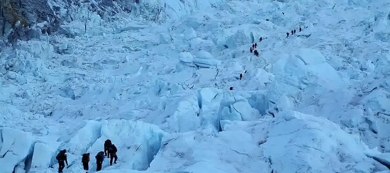 Small groups of climbers in dark clothing walking up through a broken field of blue-white ice blocks and crevasses in the Khumbu Icefall at dawn.