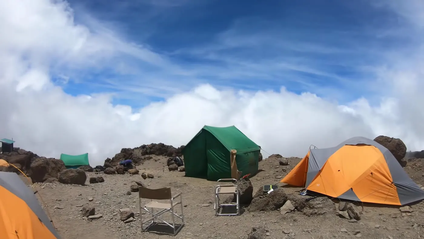 A high, rocky Kilimanjaro camp with a large green tent, an orange and gray dome tent and two folding chairs, above a bank of white cloud.