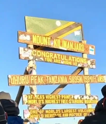 The wooden summit sign on Kilimanjaro against a blue sky, with boards reading Mount Kilimanjaro, Congratulations, Uhuru Peak Tanzania 5895 m and Africa's highest point.